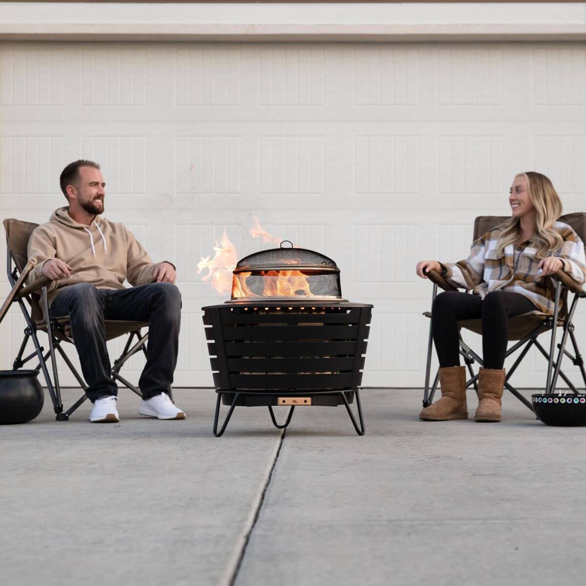 Two people relax in chairs around a TIKI® fire pit on a driveway, enjoying a warm outdoor gathering.