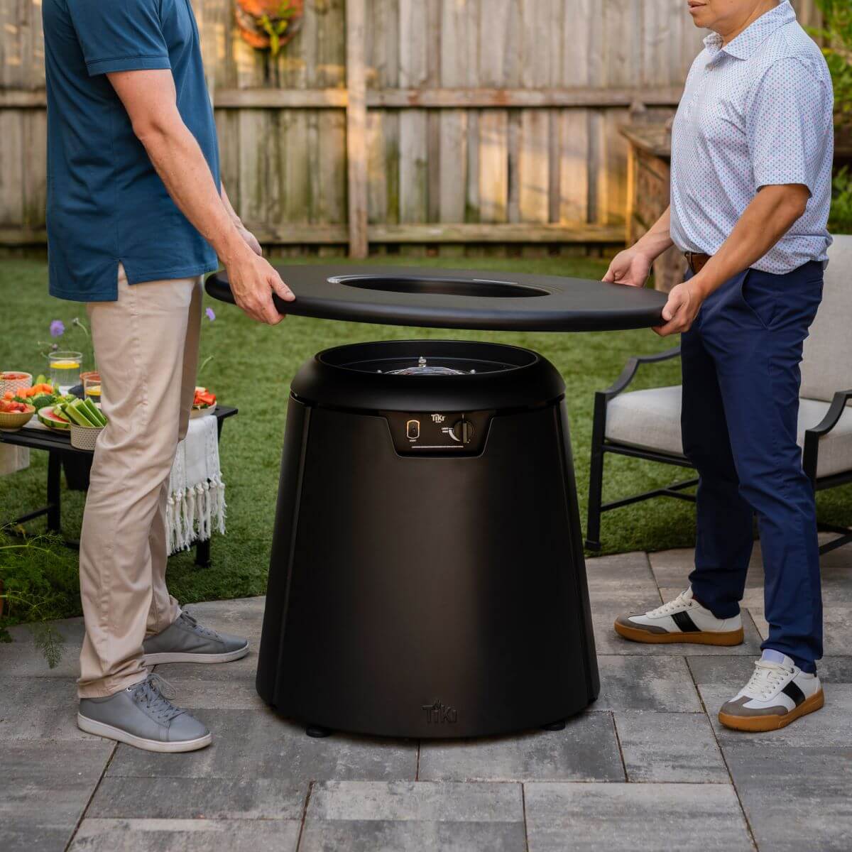 Two people lift the lid off a TIKI fire pit in a backyard patio setting, preparing for outdoor entertaining.