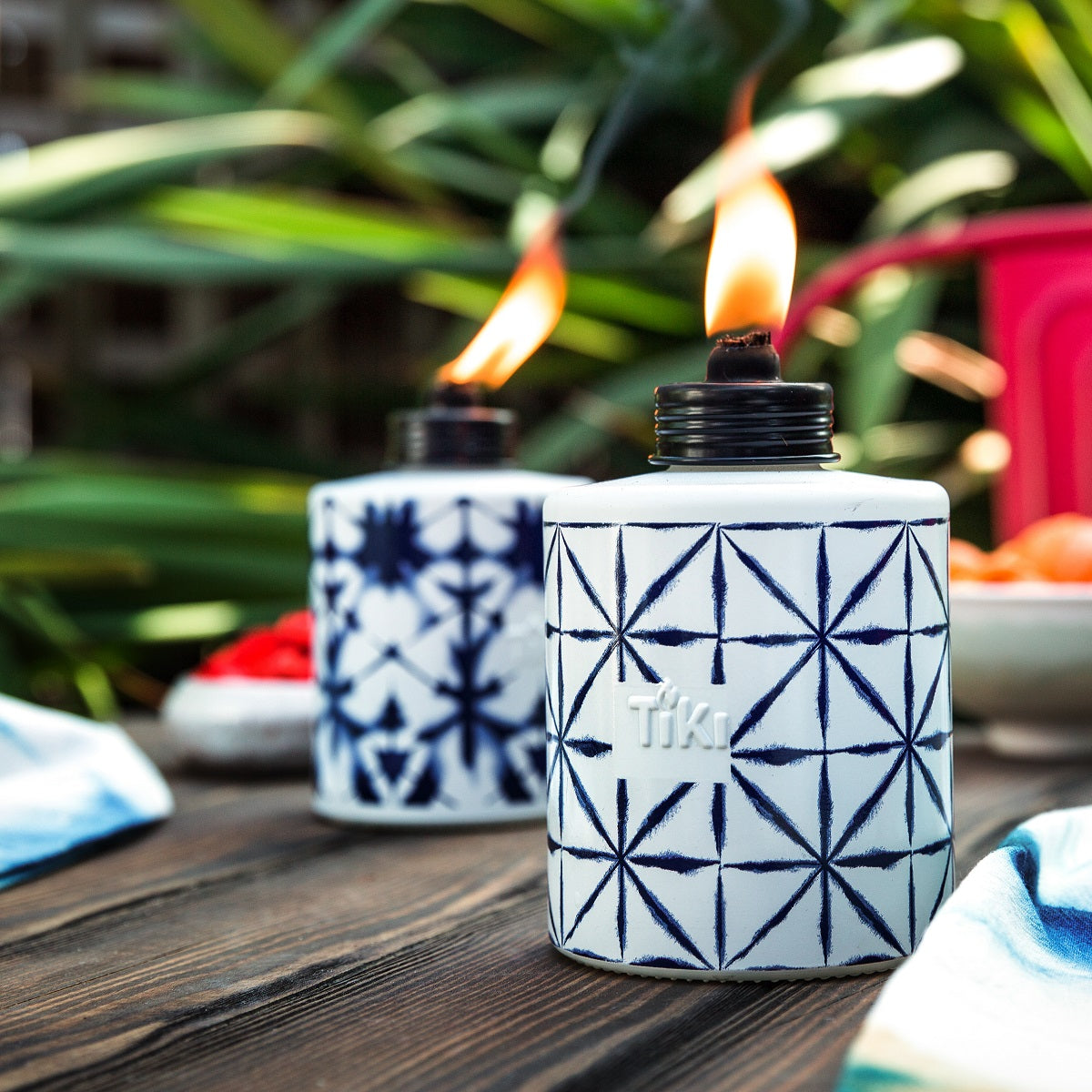 Two patterned TIKI table torches with blue geometric designs, lit outdoors on a wooden surface, surrounded by greenery.