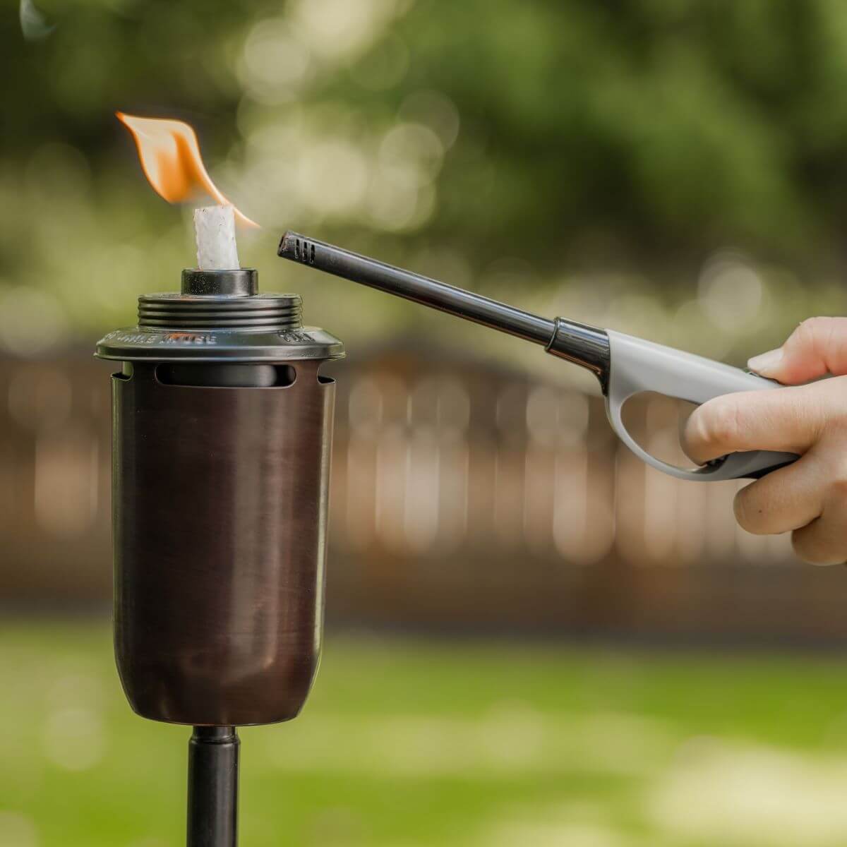 A hand lights a TIKI Brand torch outdoors using a long lighter; flame burns on the torch’s wick.