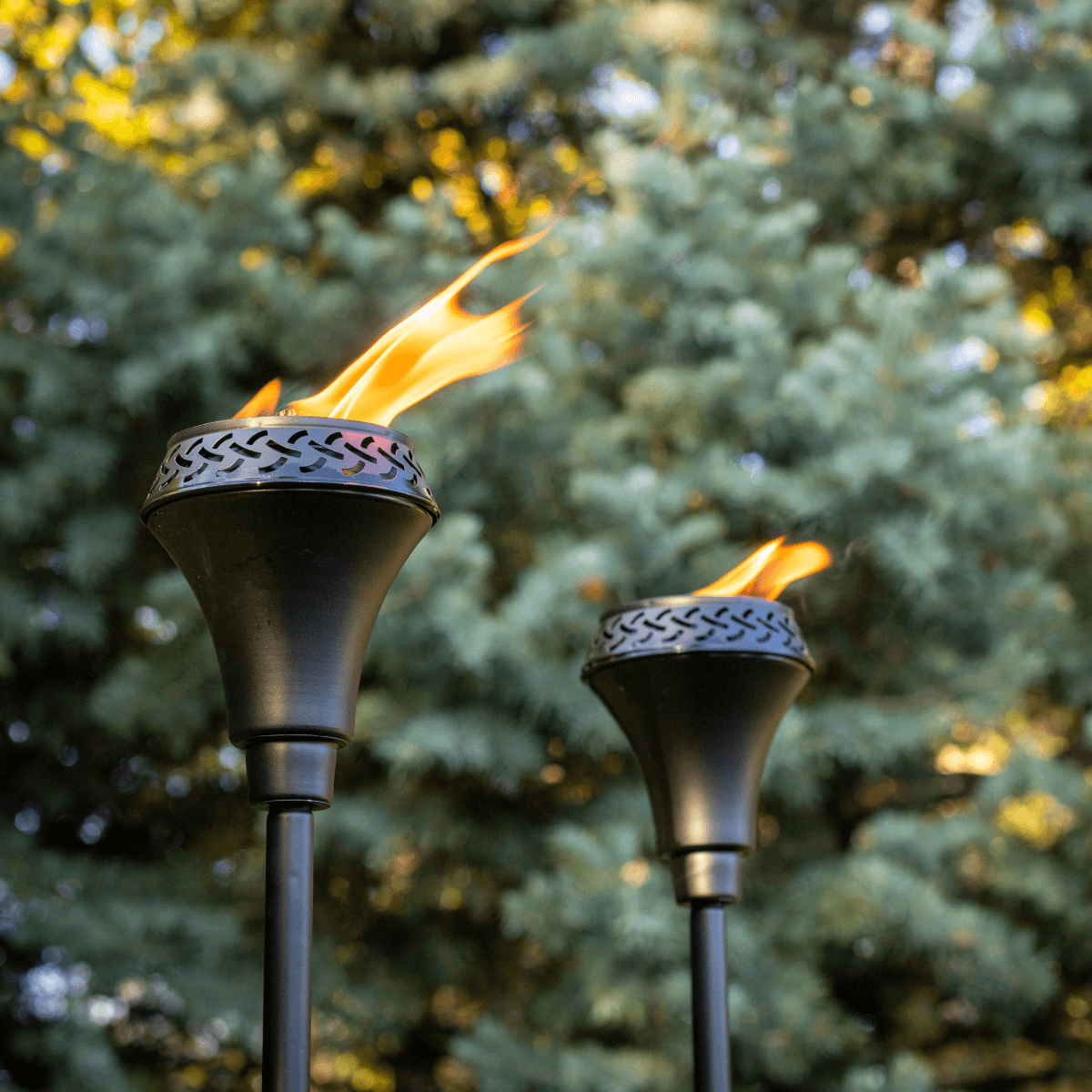 Two black TIKI® torches with flames burning outdoors, set against green trees in a backyard setting.