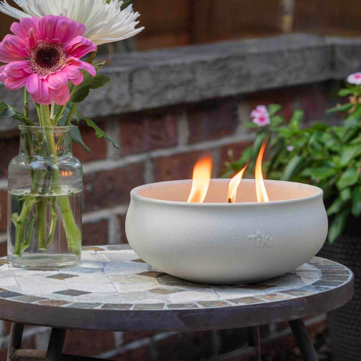 White TIKI® fire bowl with flames on a mosaic table, next to a glass vase of pink and white flowers, outdoors.