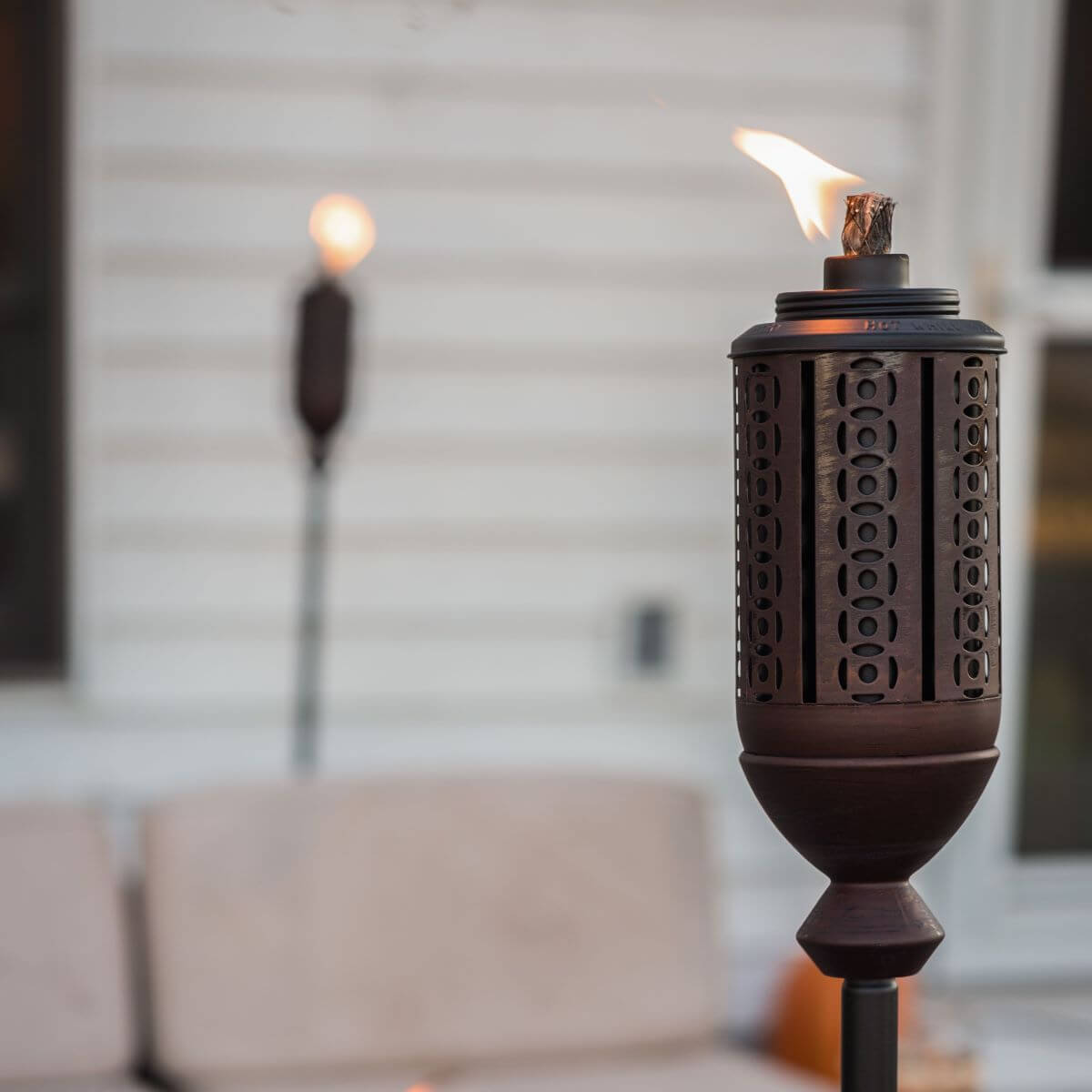 Bronze TIKI torch with patterned metal design, lit outdoors near patio seating and a white house wall.