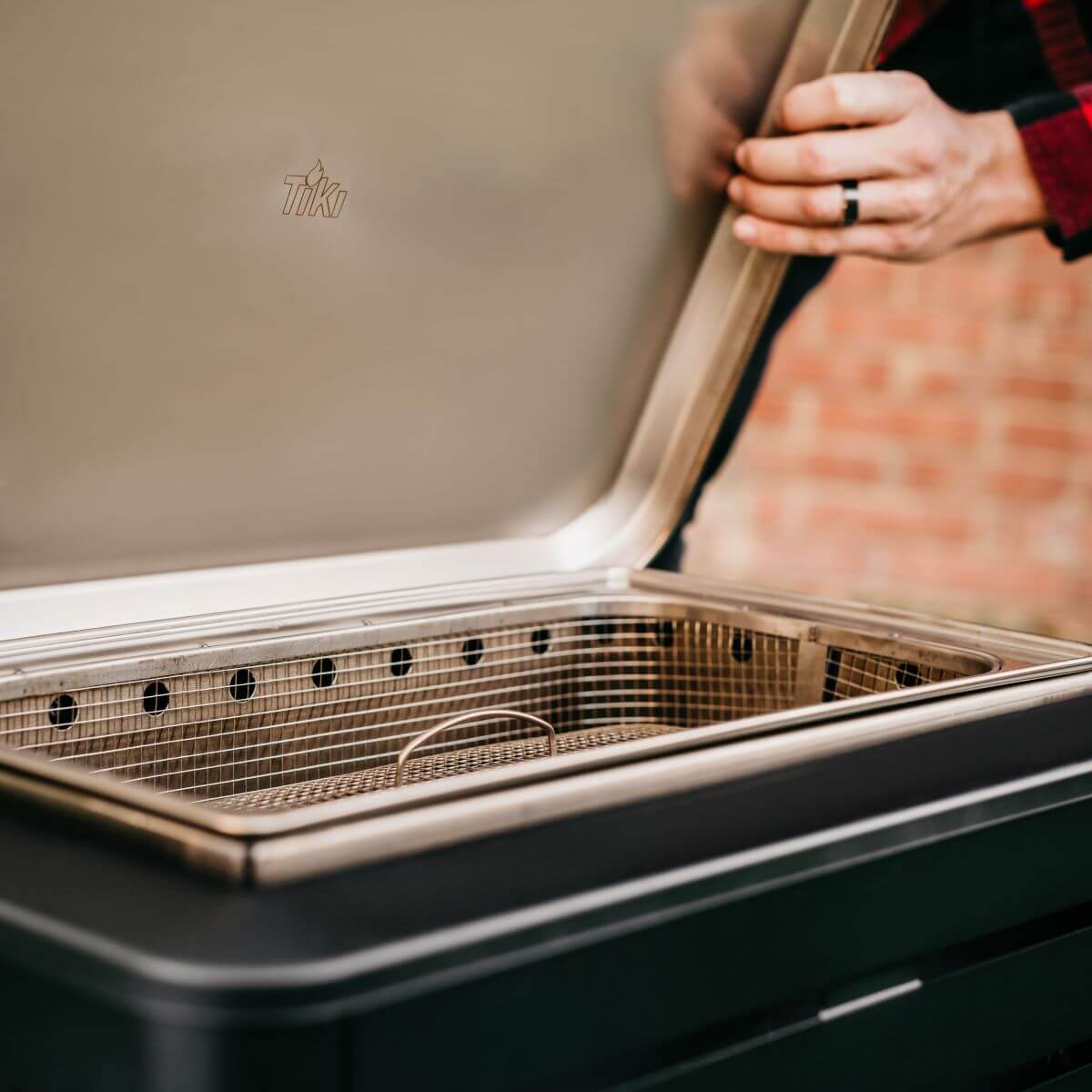 Person opening the lid of a TIKI fire pit, showing the stainless steel interior and removable ash pan.