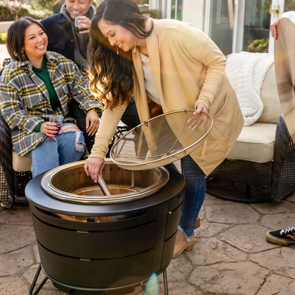 Group enjoying outdoor seating while a woman lifts the mesh cover of a TIKI® fire pit on a patio.