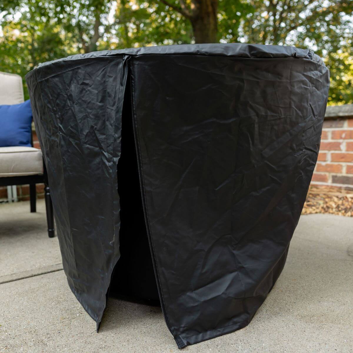Black outdoor fire pit cover with a zippered opening, protecting the fire pit on a patio near a chair and brick wall.
