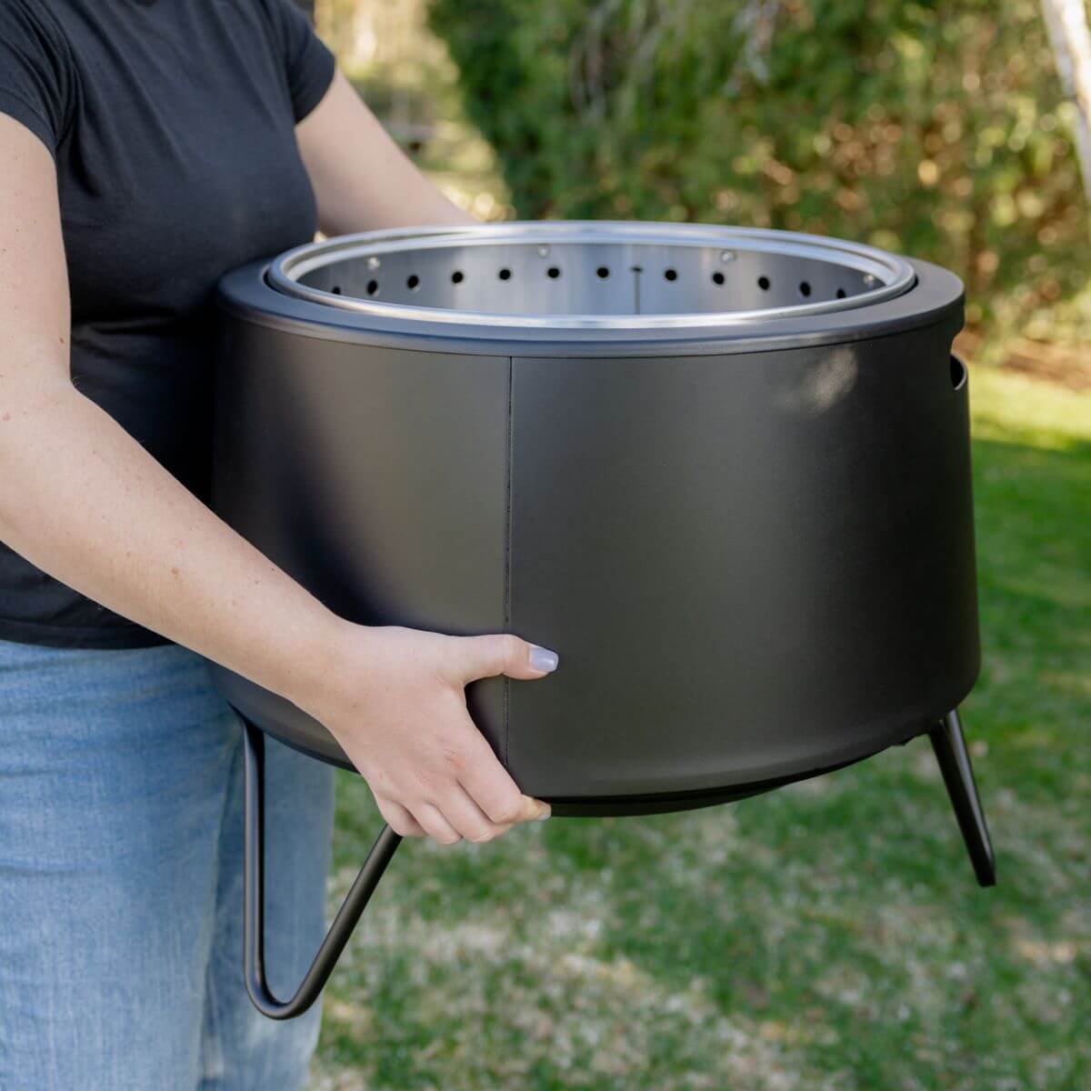 Person carrying a black TIKI® fire pit outdoors; lightweight and portable for backyard use.