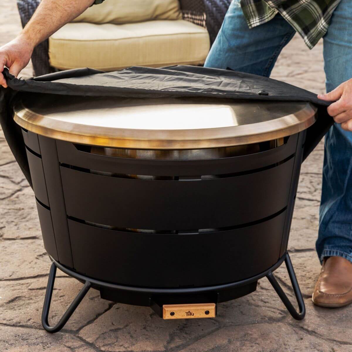 Person placing a cover over a TIKI® fire pit on a patio, with outdoor seating in the background.