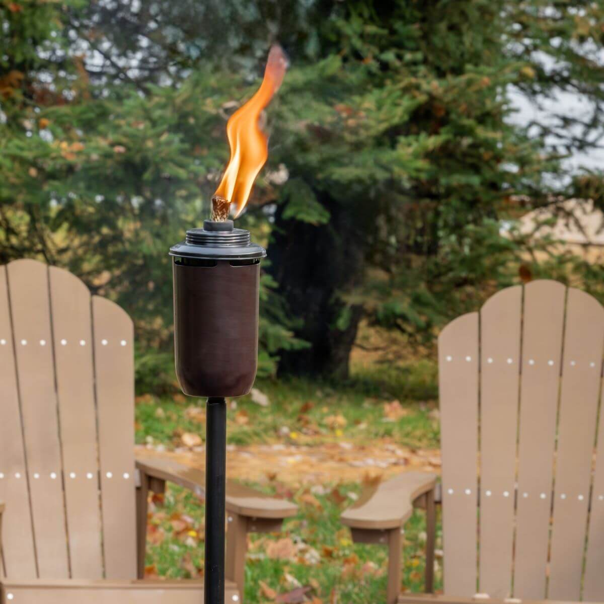A TIKI® torch with a bright flame stands in front of two Adirondack chairs in a backyard setting.
