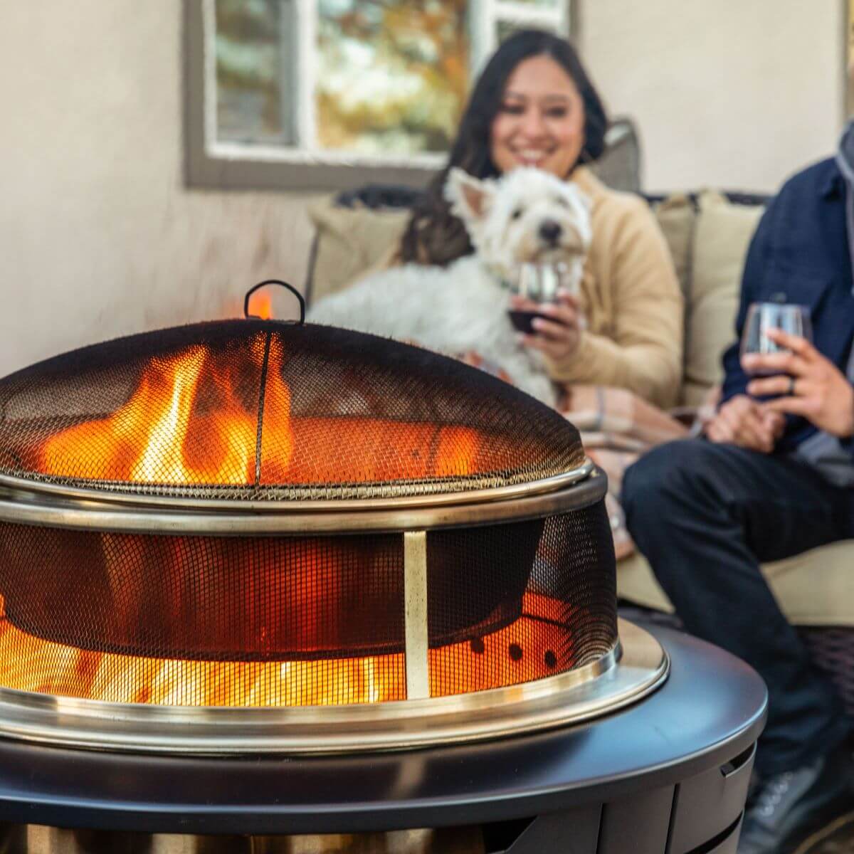 Outdoor fire pit with bright flames; people and a dog relaxing nearby, enjoying drinks on a patio.