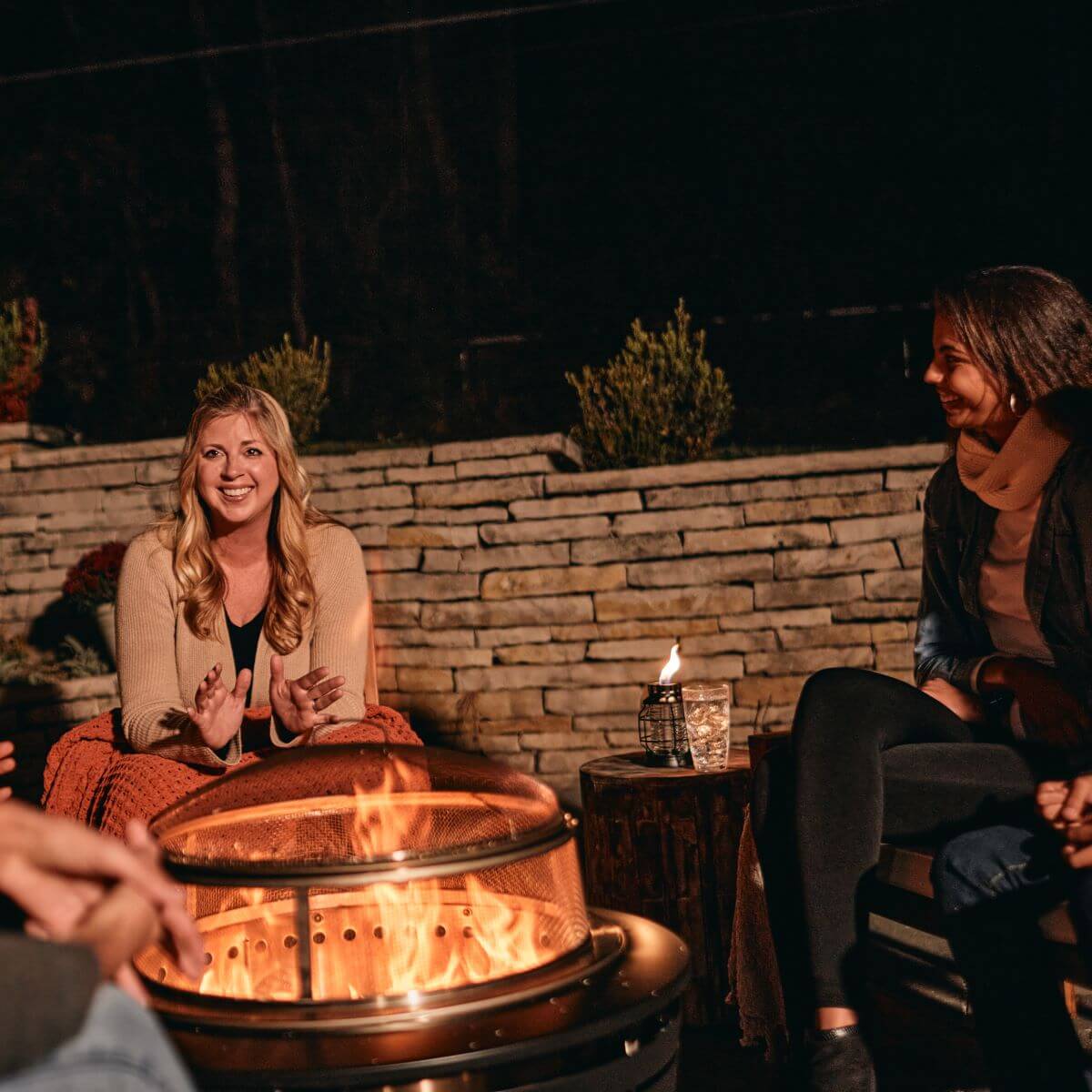 Group enjoying a cozy evening around a TIKI fire pit in a backyard with stone wall and outdoor torch.