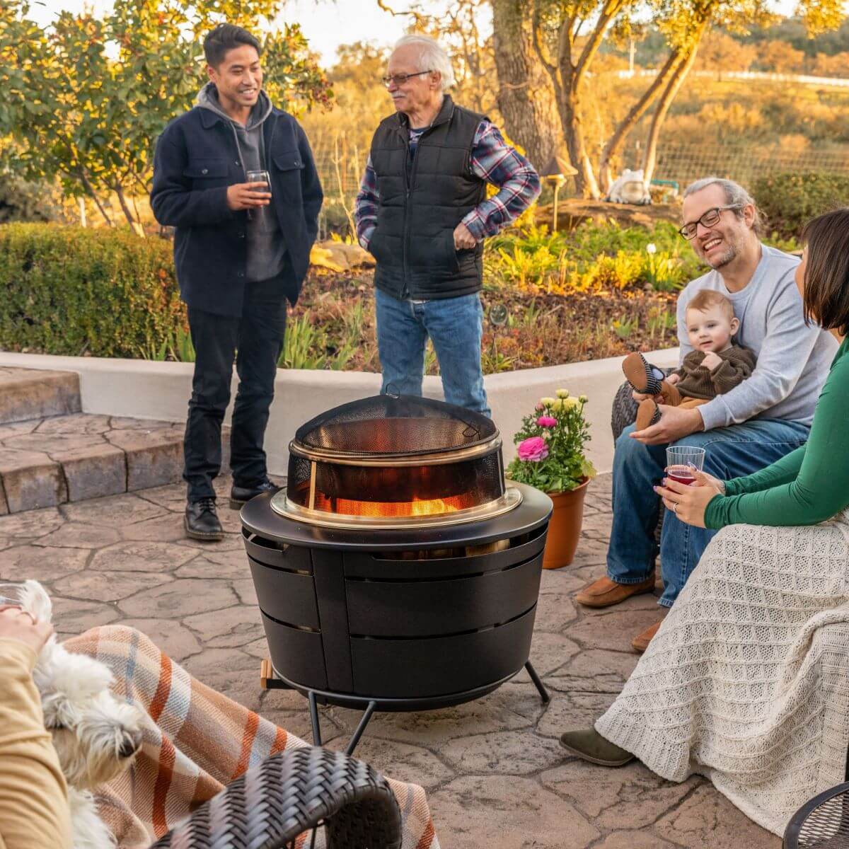 Group enjoying drinks and conversation around a TIKI Brand fire pit on a patio, with flowers and a dog nearby.