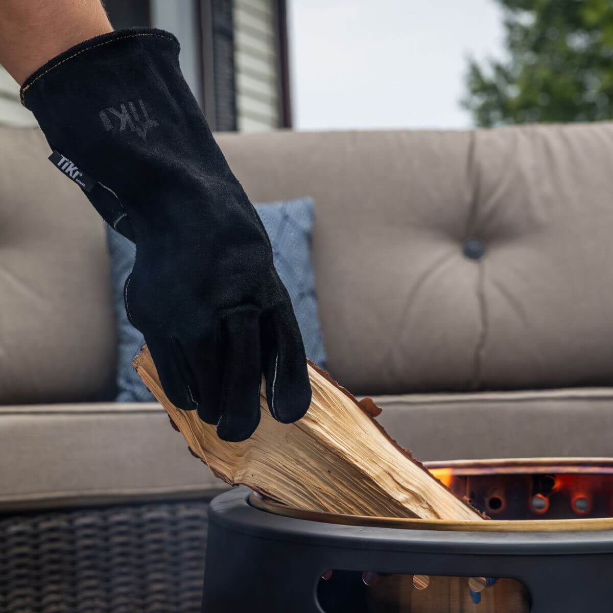 A person wearing a TIKI-branded heat-resistant glove adds firewood to a patio fire pit in an outdoor seating area.
