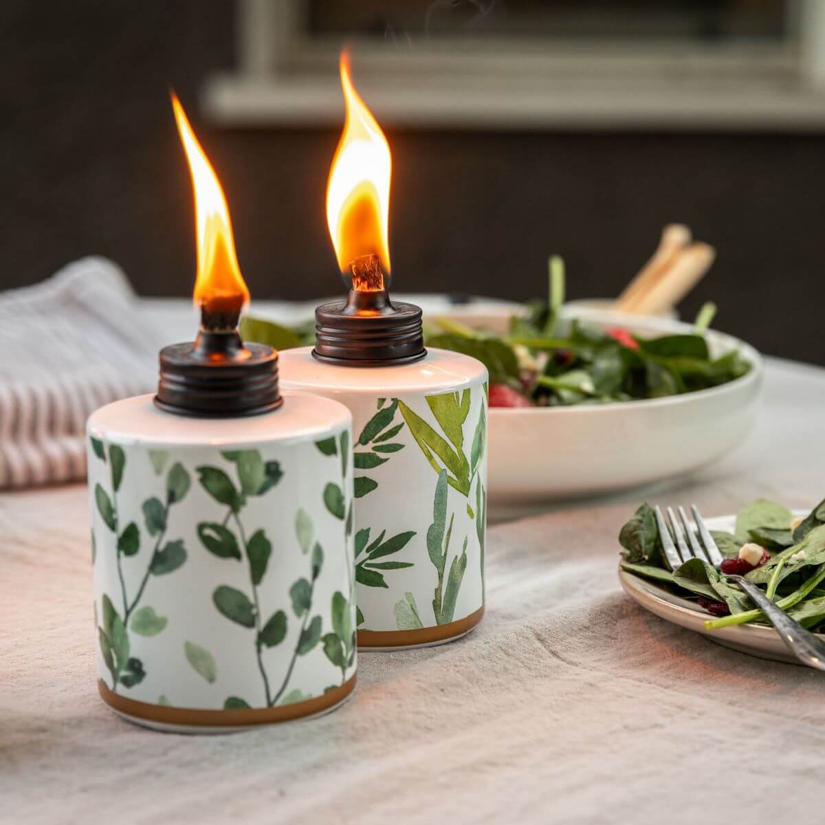 Two botanical-patterned TIKI tabletop torches with flames, set on an outdoor dining table next to a salad plate.