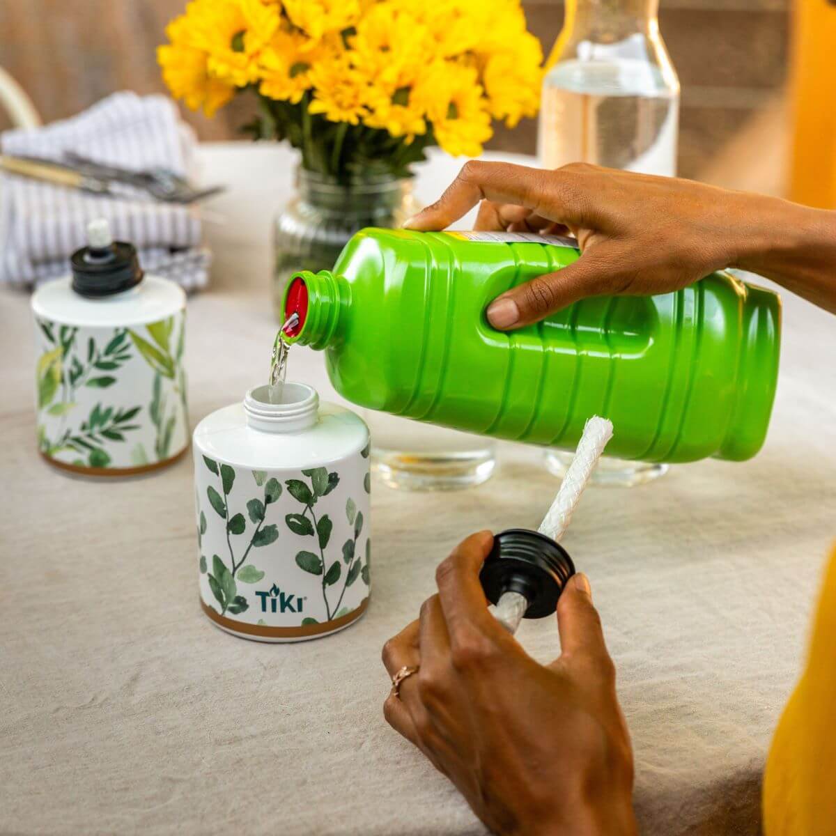 A person pours fuel into a TIKI Brand tabletop torch with a botanical design, preparing it for outdoor use.