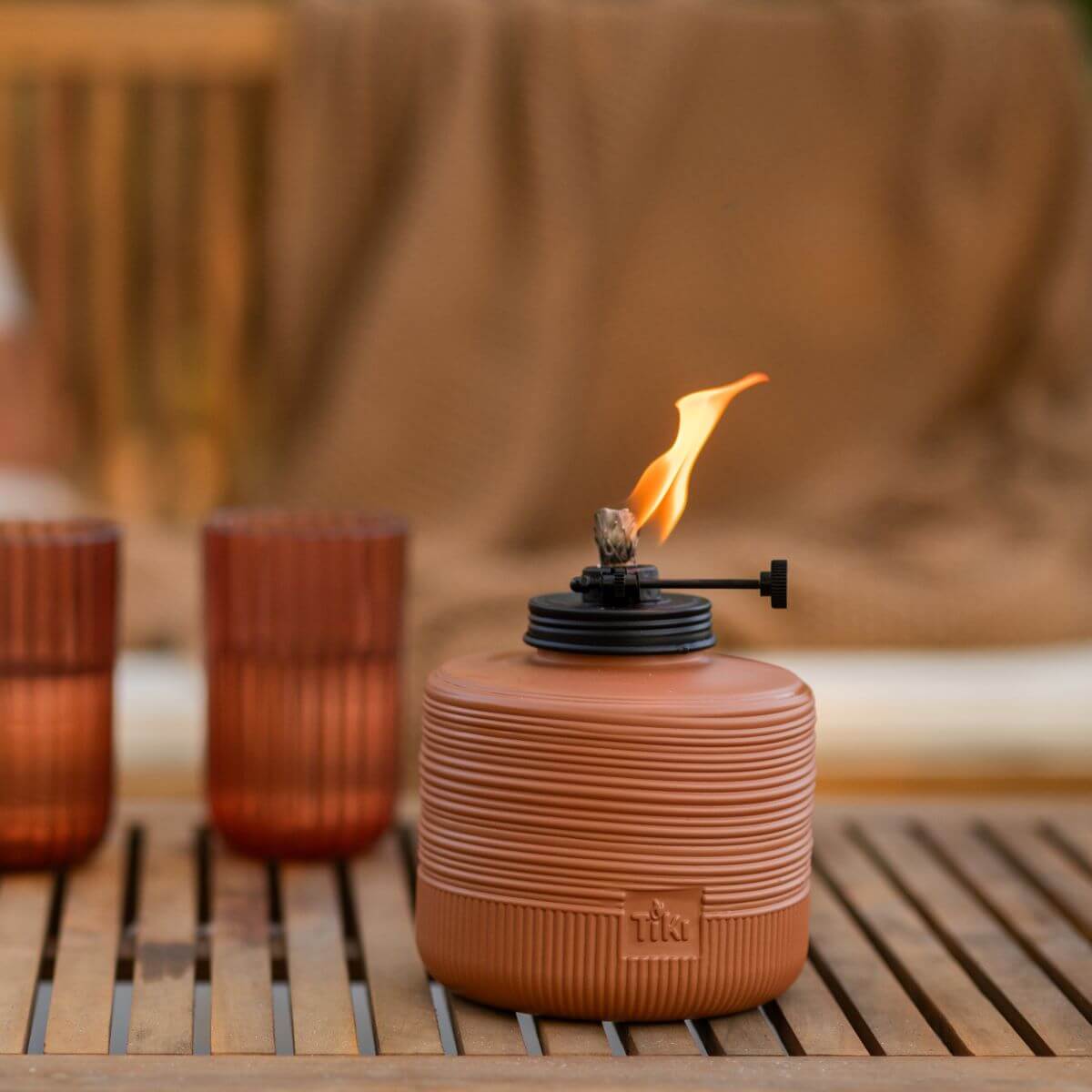Terracotta TIKI tabletop fire pit with flame, placed on a wooden outdoor table beside two amber glasses.