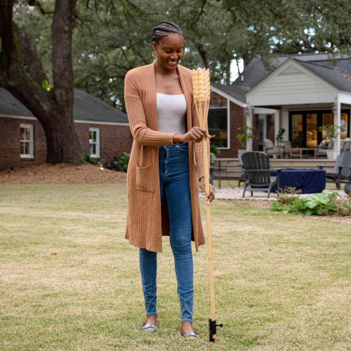 Person standing in a backyard holding a bamboo TIKI torch, with patio seating and a house in the background.
