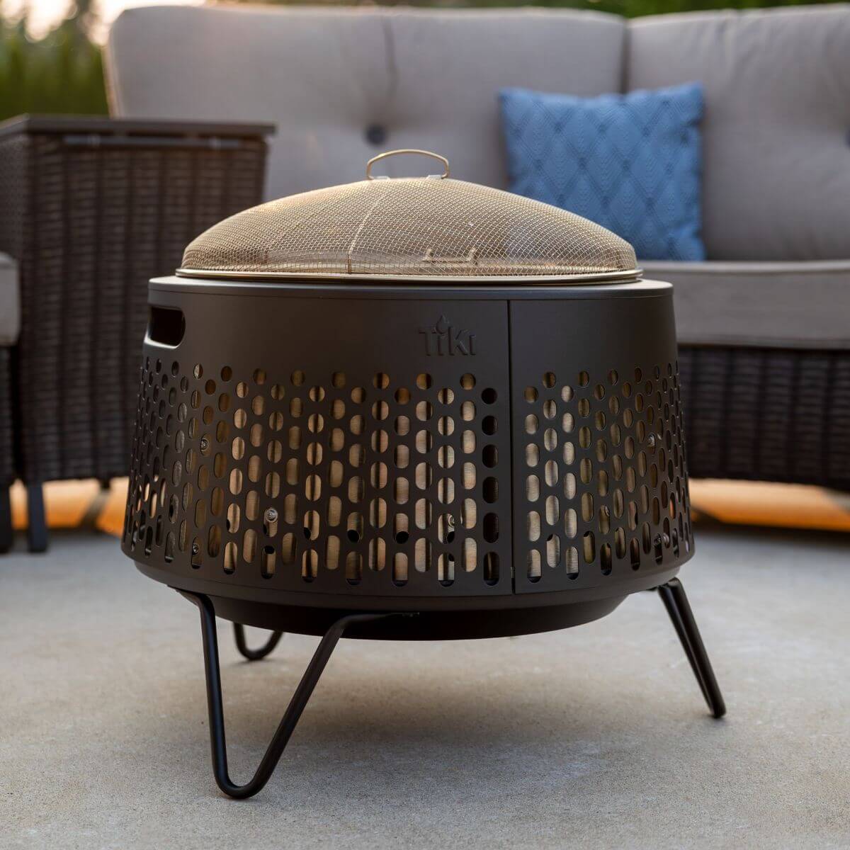 Black TIKI fire pit with mesh lid on a patio, placed in front of outdoor wicker furniture and a blue cushion.