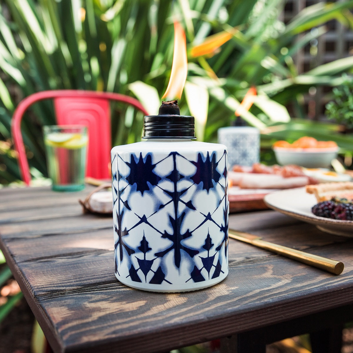 A blue and white patterned TIKI® table torch with a flame, set on an outdoor wooden table with food and drinks.