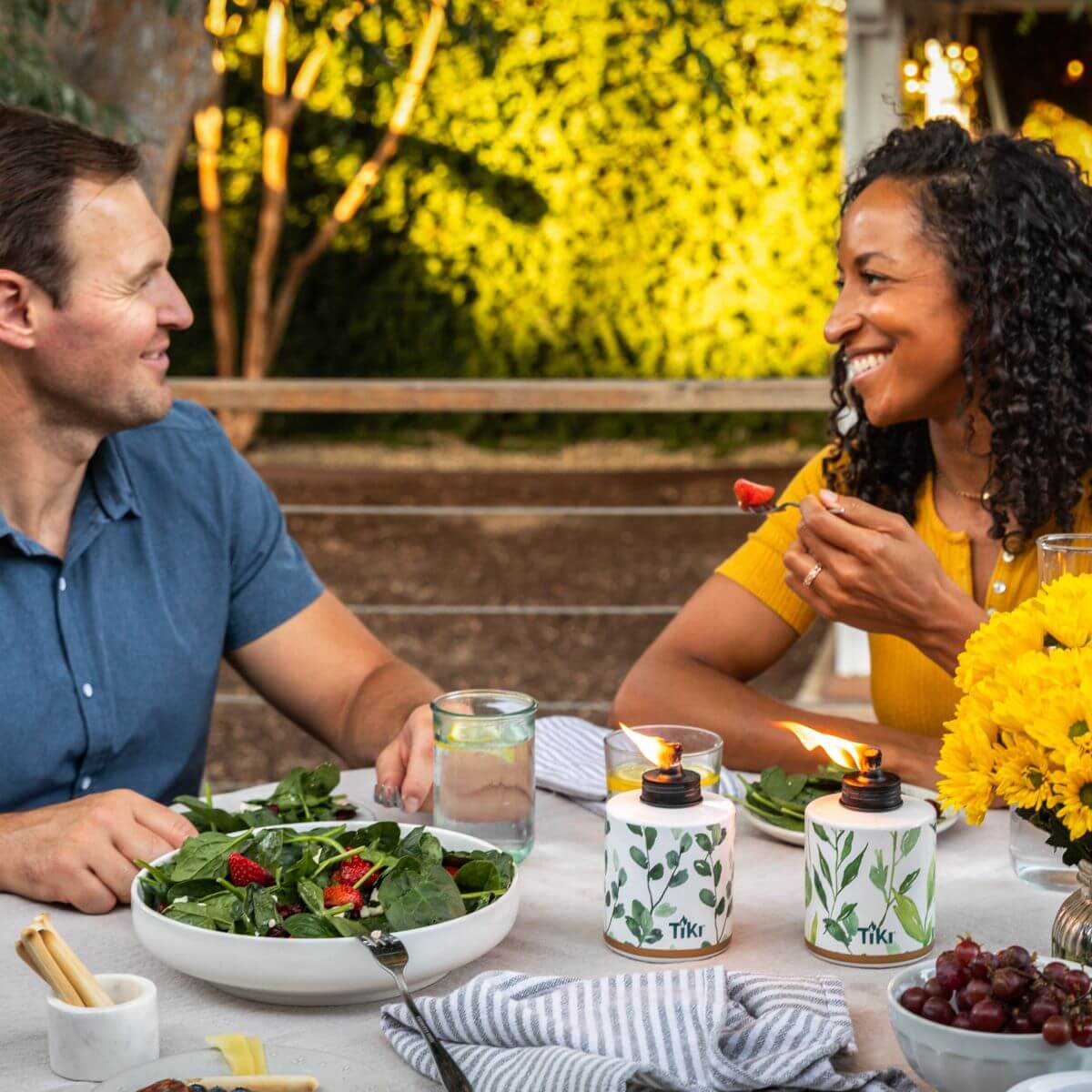 Outdoor dining scene with TIKI Brand tabletop torches, salad, flowers, and drinks on a patio table.