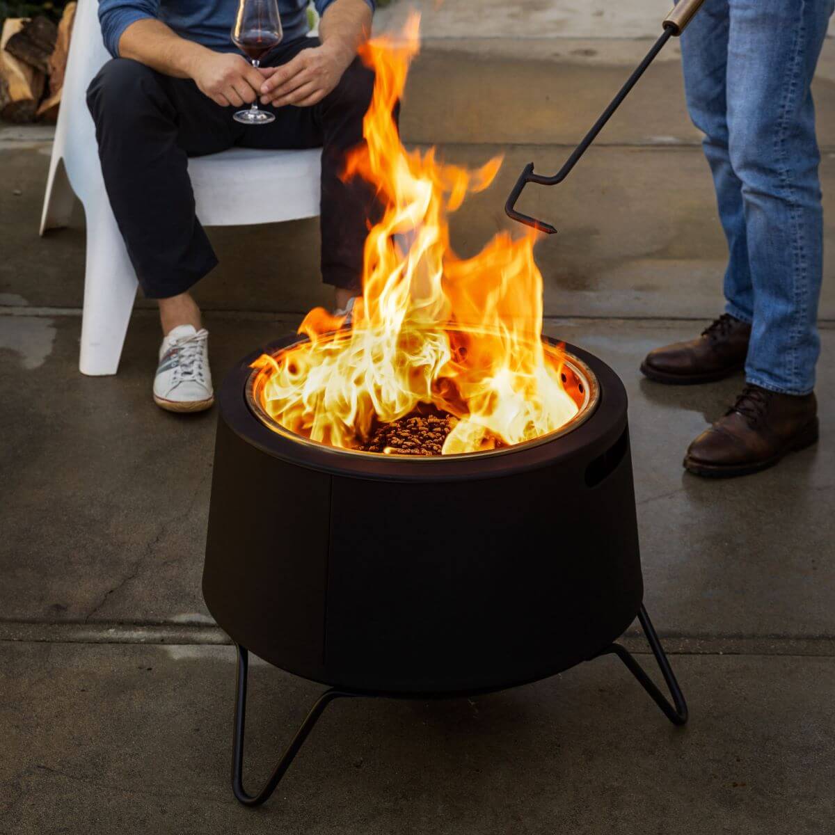 Two people enjoy a TIKI® fire pit on a patio; one sits with a drink, the other tends the fire with a poker.