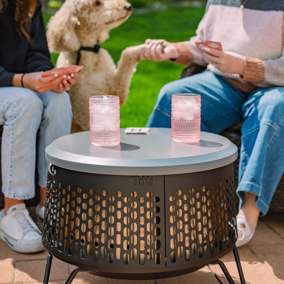 Two people play cards outdoors beside a TIKI fire pit table, with drinks and a dog joining them on a sunny patio.