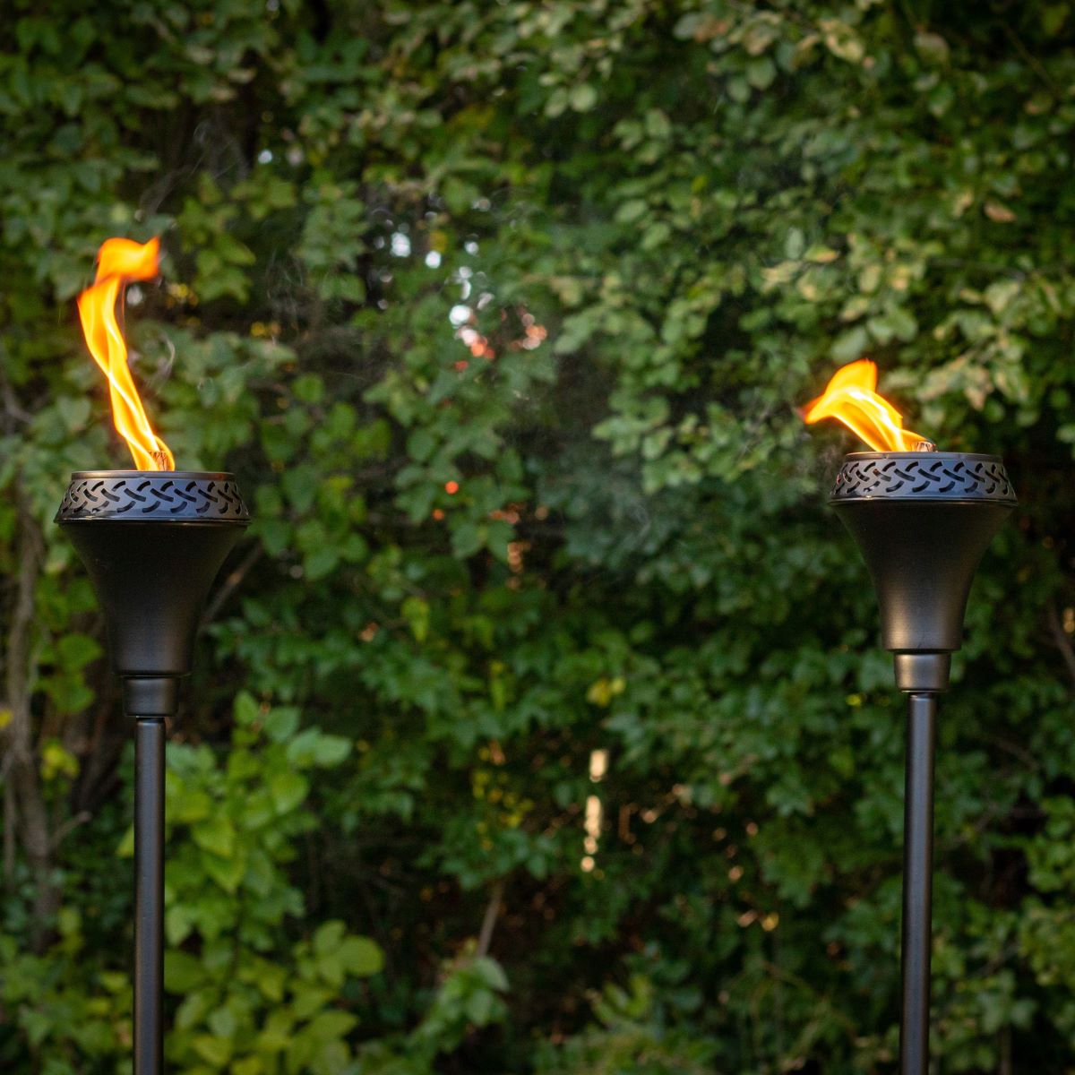 Two black TIKI® torches with bright flames, set outdoors against lush green foliage.