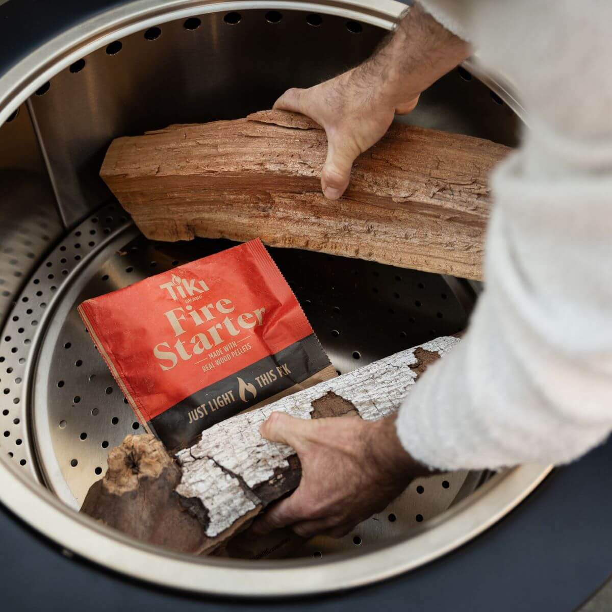 A person places logs and a TIKI Fire Starter packet into a fire pit, preparing to safely start a wood fire outdoors.