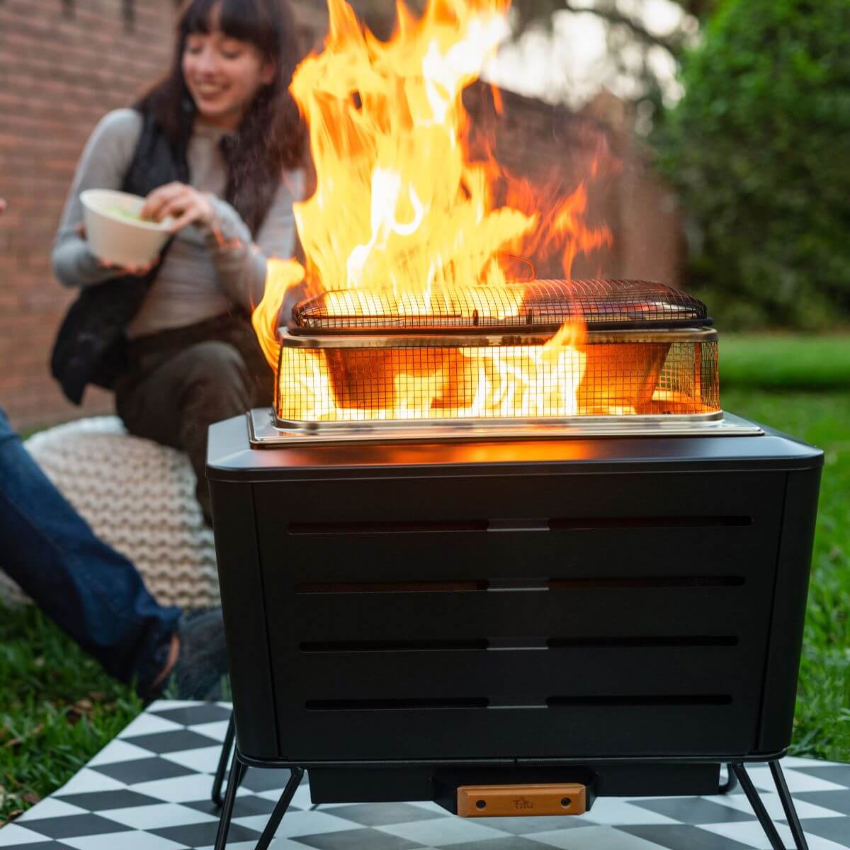 TIKI Brand fire pit burning brightly outdoors, with a person enjoying food nearby on a grassy lawn.