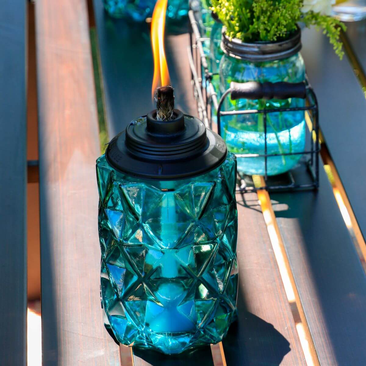 Blue glass TIKI® torch with geometric pattern lit on an outdoor table, adding ambiance to a patio setting.