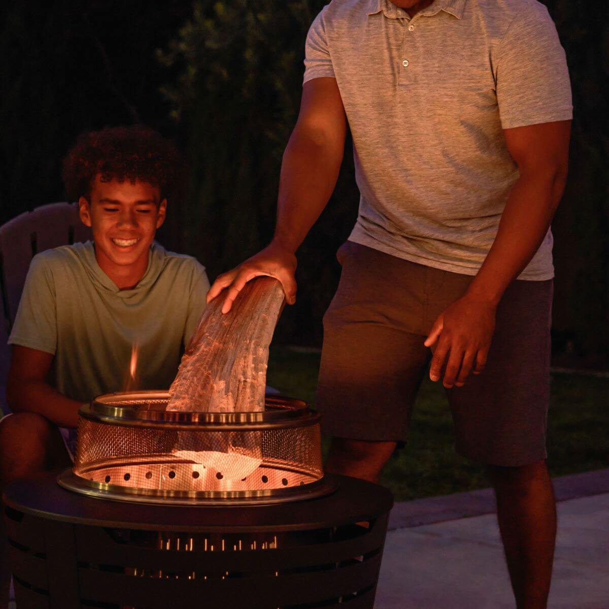 Two people enjoy an evening outdoors, adding wood to a TIKI® fire pit for a cozy backyard gathering.