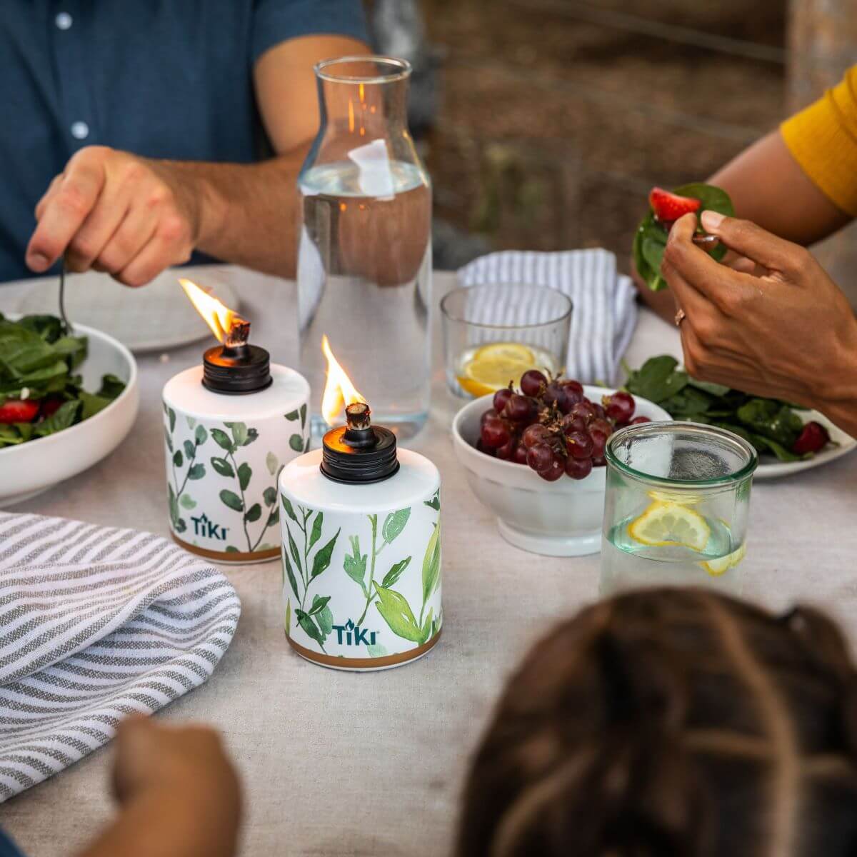 Outdoor dining table with TIKI tabletop torches, salad, grapes, and drinks; people enjoying a meal together.