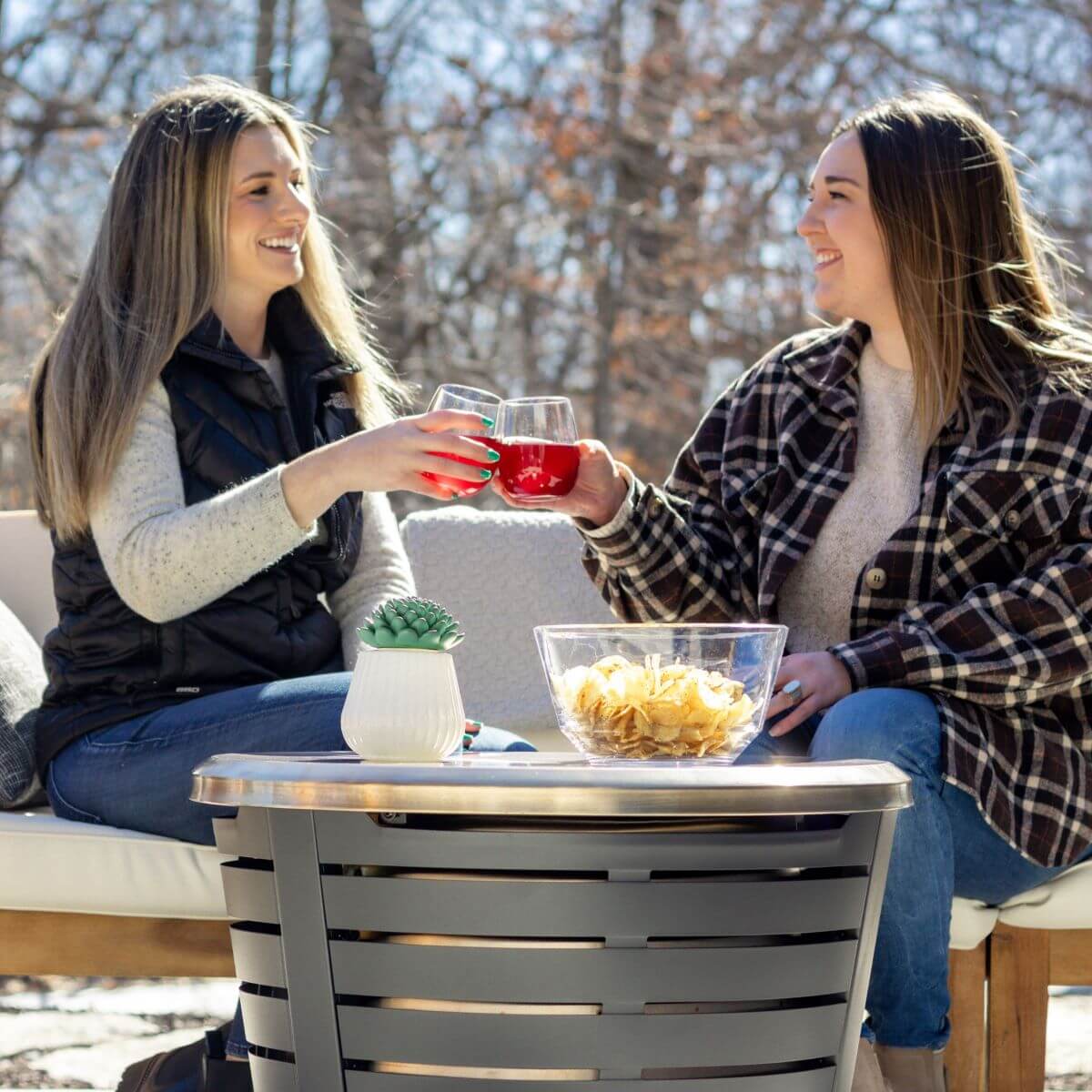 Two women enjoy drinks and snacks outdoors beside a TIKI® fire pit table, with trees in the background.