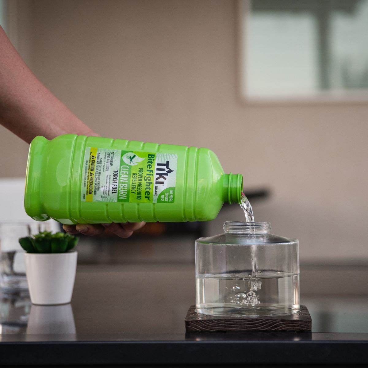 A hand pours TIKI BiteFighter torch fuel into a clear tabletop torch on an outdoor patio table.