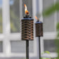 Brown woven TIKI® torch with flame, set outdoors near a window, ideal for backyard ambiance and lighting.