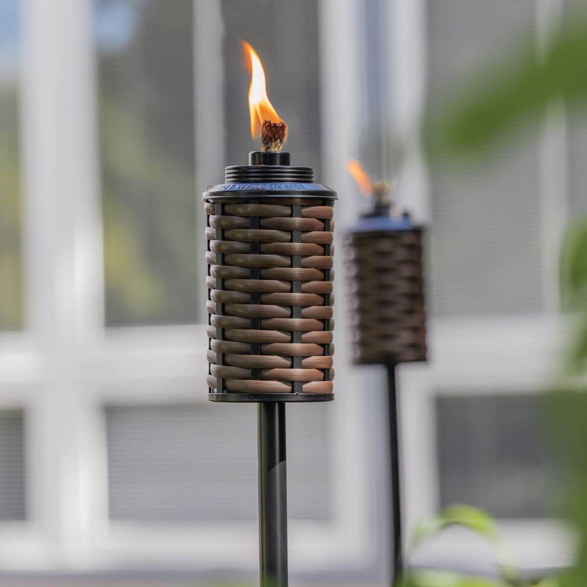 Brown woven TIKI® torch with flame, set outdoors near a window, ideal for backyard ambiance and lighting.