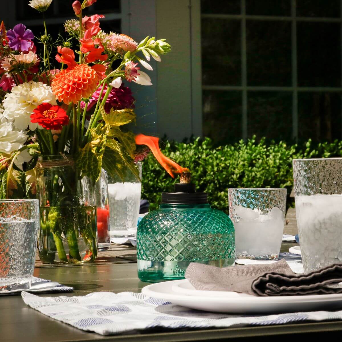 A teal glass TIKI® table torch burns on an outdoor dining table set with flowers, glasses, and plates.