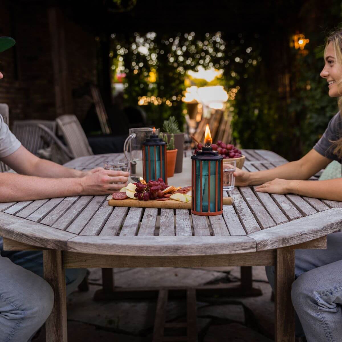 Two people enjoy snacks outdoors at a wooden table with TIKI® Brand lanterns, creating a cozy, inviting atmosphere.