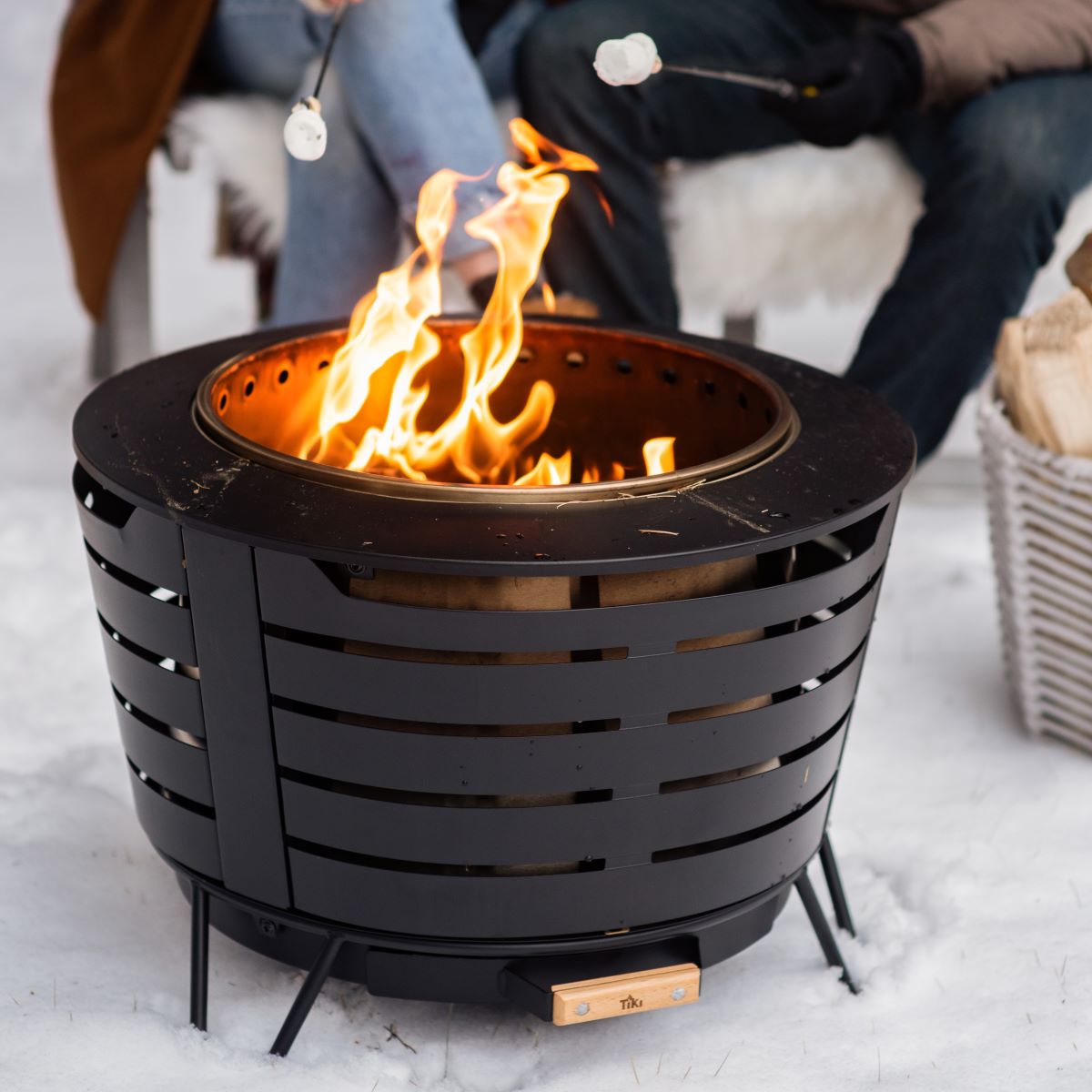 Round black metal fire pit with open flames, set on snow. Two people roast marshmallows nearby.