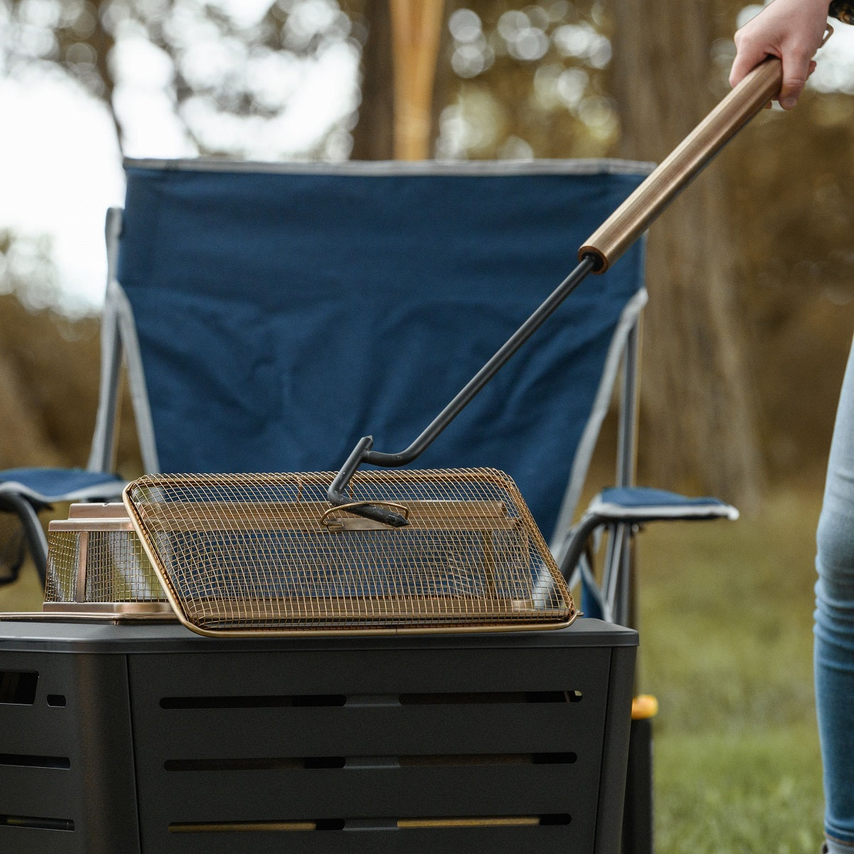 Person using a long-handled tool to lift a mesh spark screen from a TIKI fire pit outdoors.