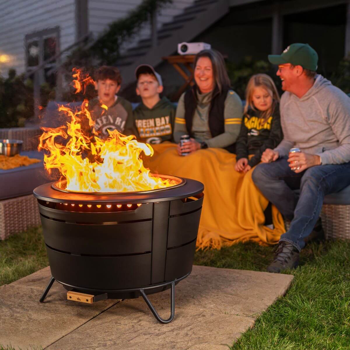 Group enjoys a cozy evening outdoors around a TIKI® fire pit, wrapped in blankets and watching a game together.