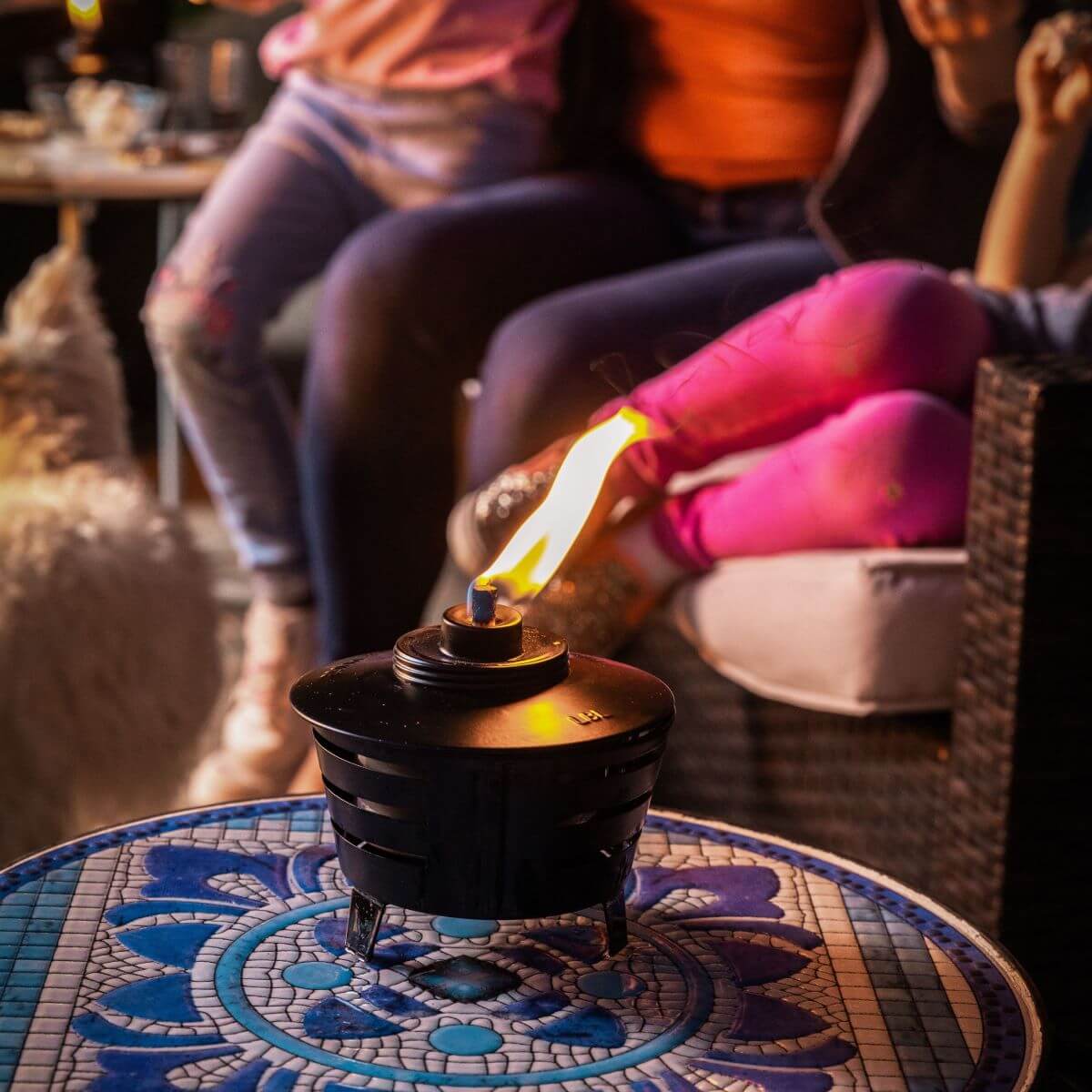A TIKI tabletop fire pit burns on a mosaic table, with people relaxing in the background.