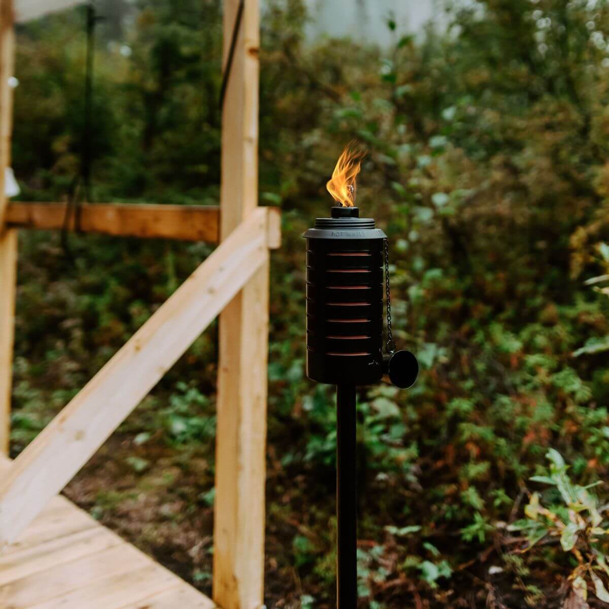 Black TIKI torch with flame lit, placed outdoors near wooden deck and greenery.