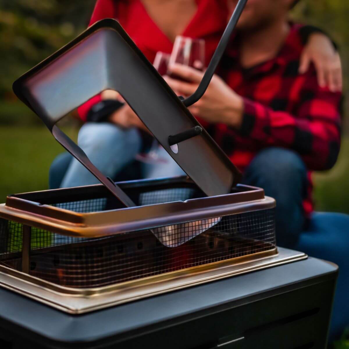 Close-up of a TIKI® fire pit with a couple in the background enjoying drinks outdoors.