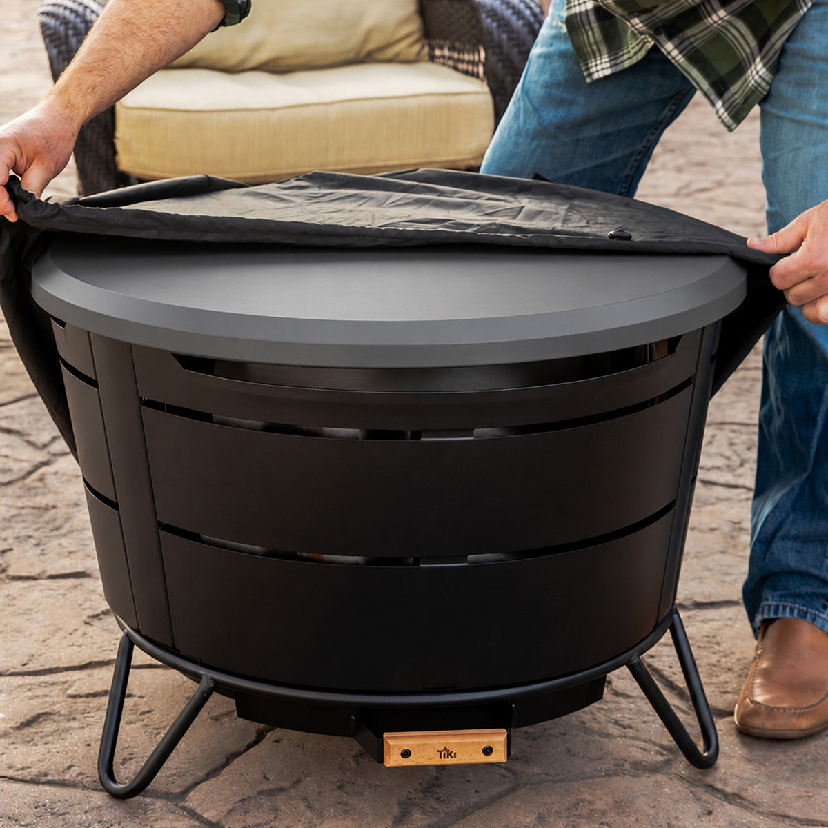 TIKI Brand Reunion Tabletop Cover Gray, being placed on fire pit outdoors by two people