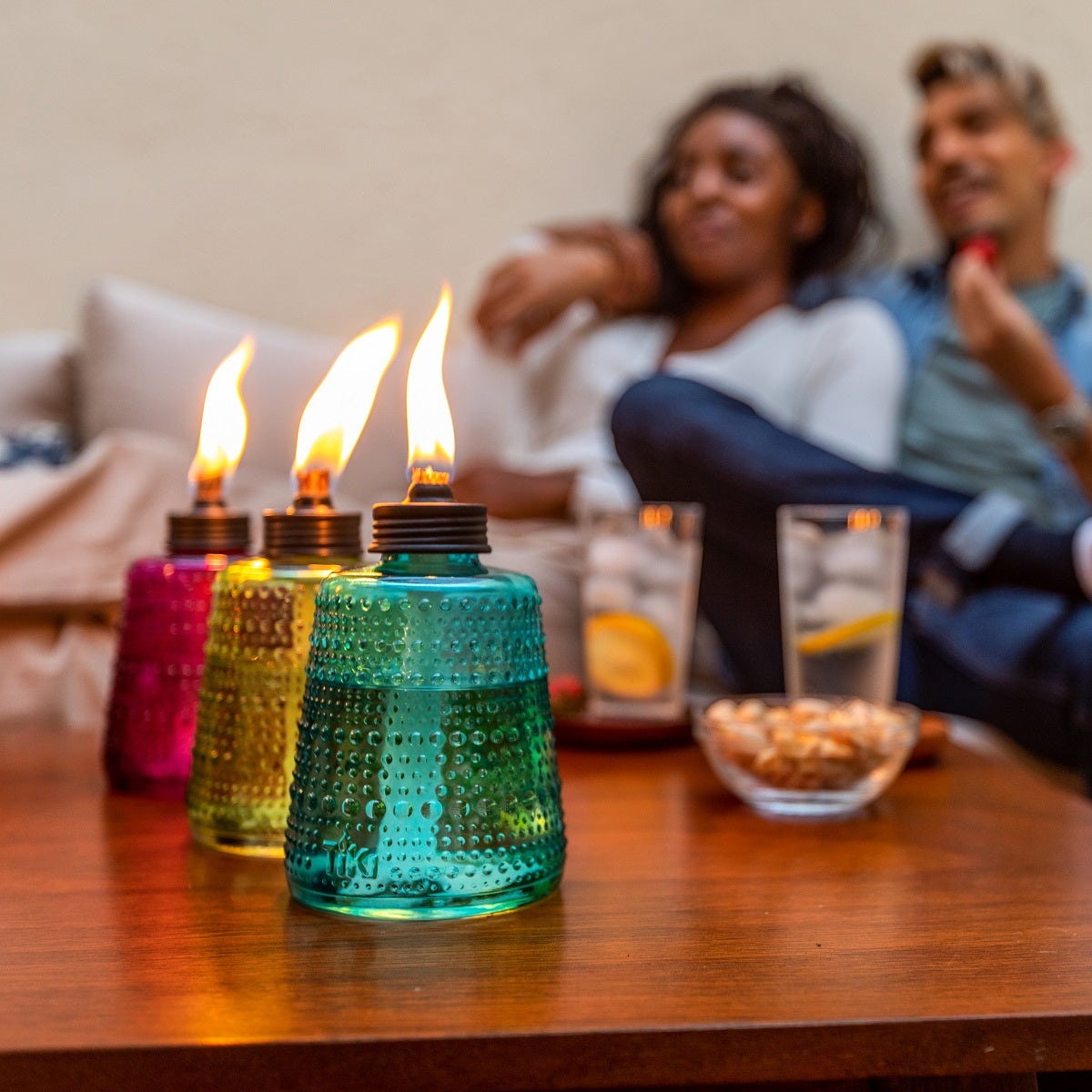 Three colorful TIKI® Brand table torches lit on a patio table, with drinks and snacks in the background.
