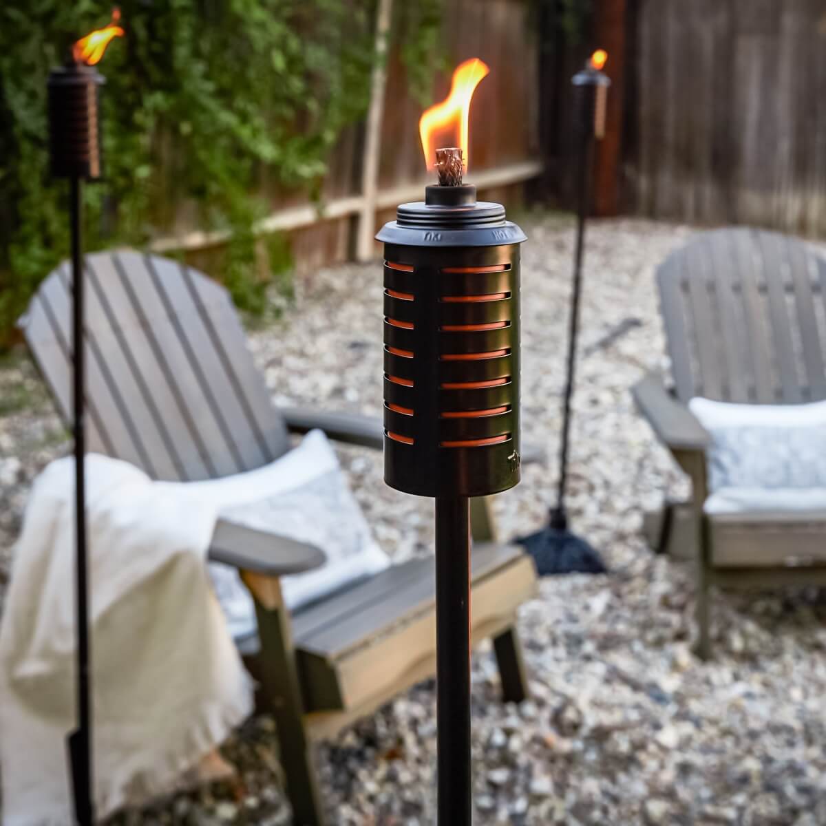 Black metal TIKI torch lit in a backyard with Adirondack chairs and gravel, creating a cozy outdoor seating area.