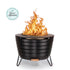 Round black metal fire pit with visible flames, sturdy legs, and a modern design. "The Spruce Approved" badge shown.