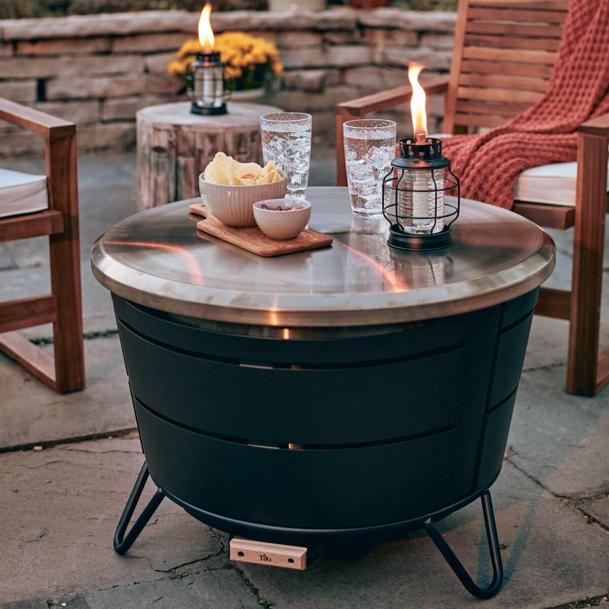 Modern patio with a TIKI® fire pit table, snacks, drinks, and a TIKI® lantern, surrounded by wooden chairs.