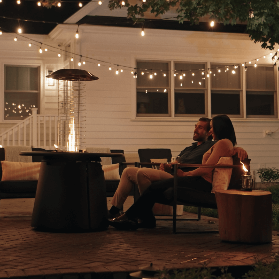 Couple relaxing on a patio at night with a TIKI® Brand fire pit and string lights creating a cozy outdoor atmosphere.